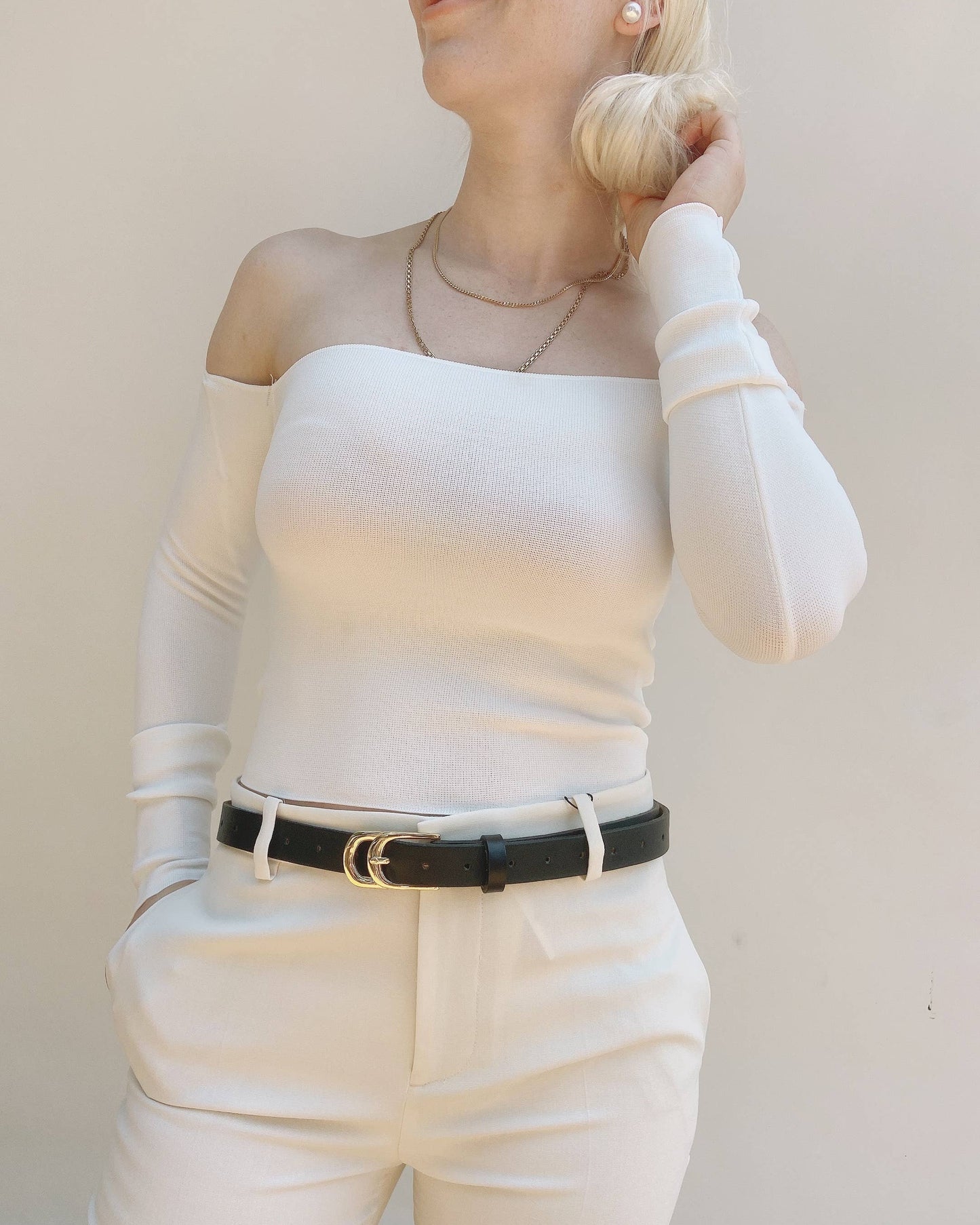 Person wearing a white long-sleeve top and beige pants with a black belt against a plain background