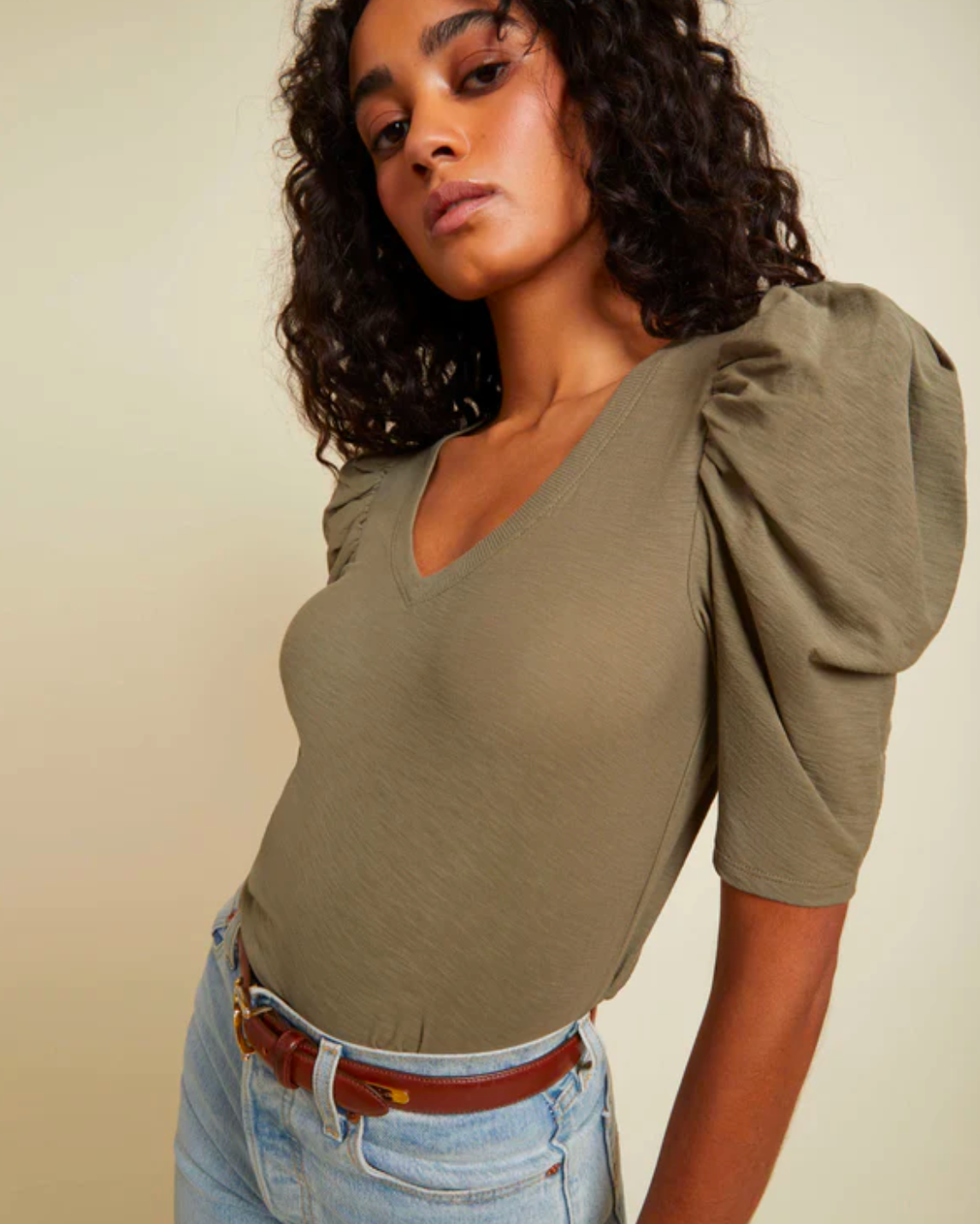 Model wearing Nation LTD Jillian Bold Shoulder V Neck in Ivy color wearing light jeans and a brown belt on a yellow background