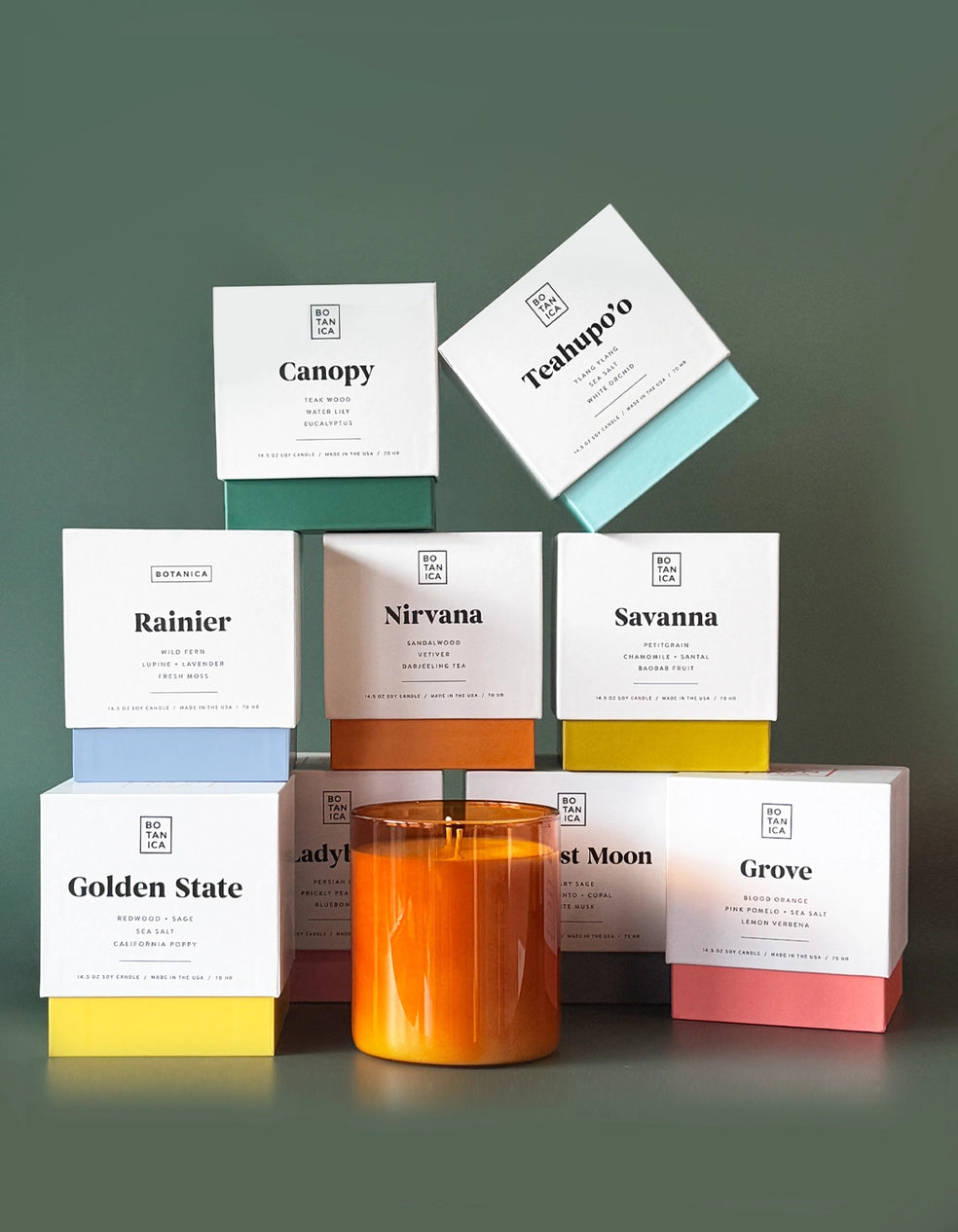 Photo of various Botanica scented candle boxes with a candle in front of a green backdrop