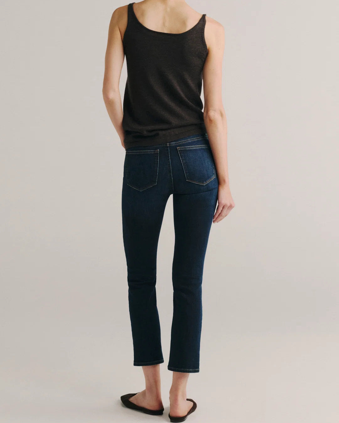 A model shown from the back wearing dark blue cropped skinny jeans, a black tank top, and black flat sandals against a light gray studio background.