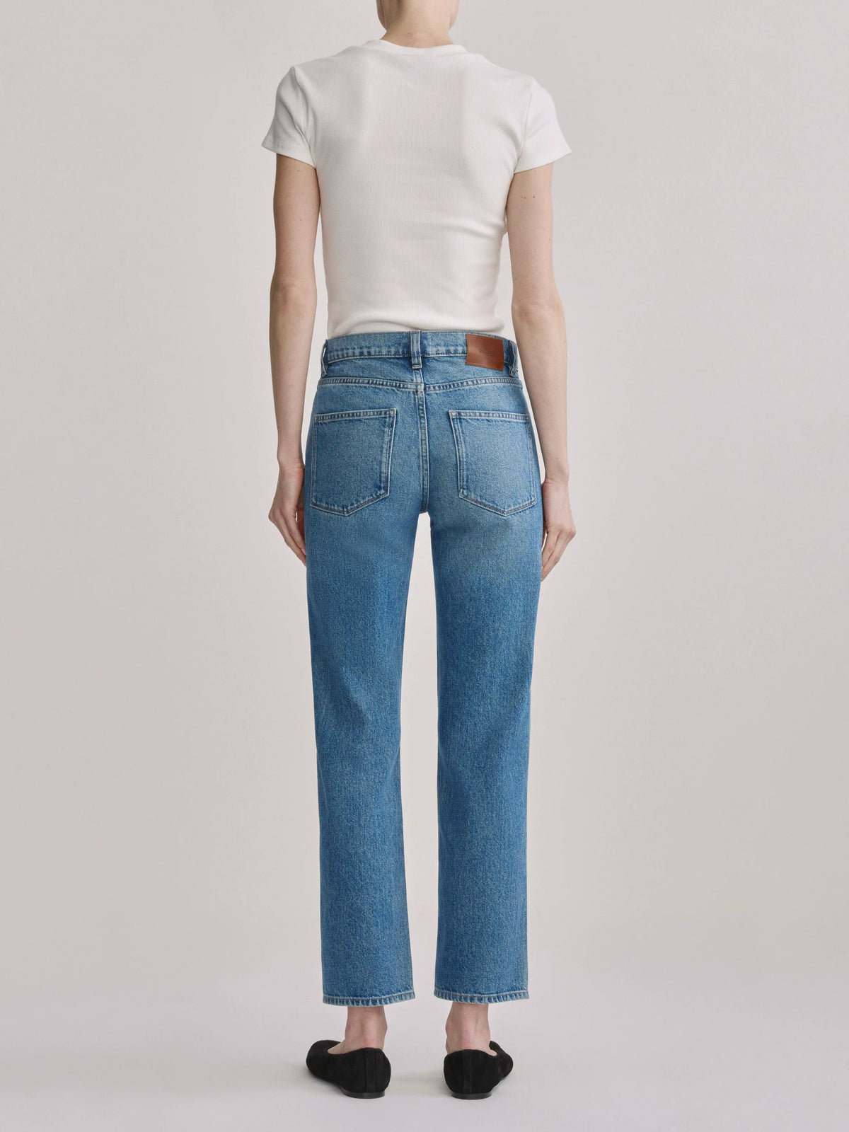 A model shown from the back wearing light-wash blue straight-leg jeans, a white t-shirt, and black flat shoes against a light gray studio background.