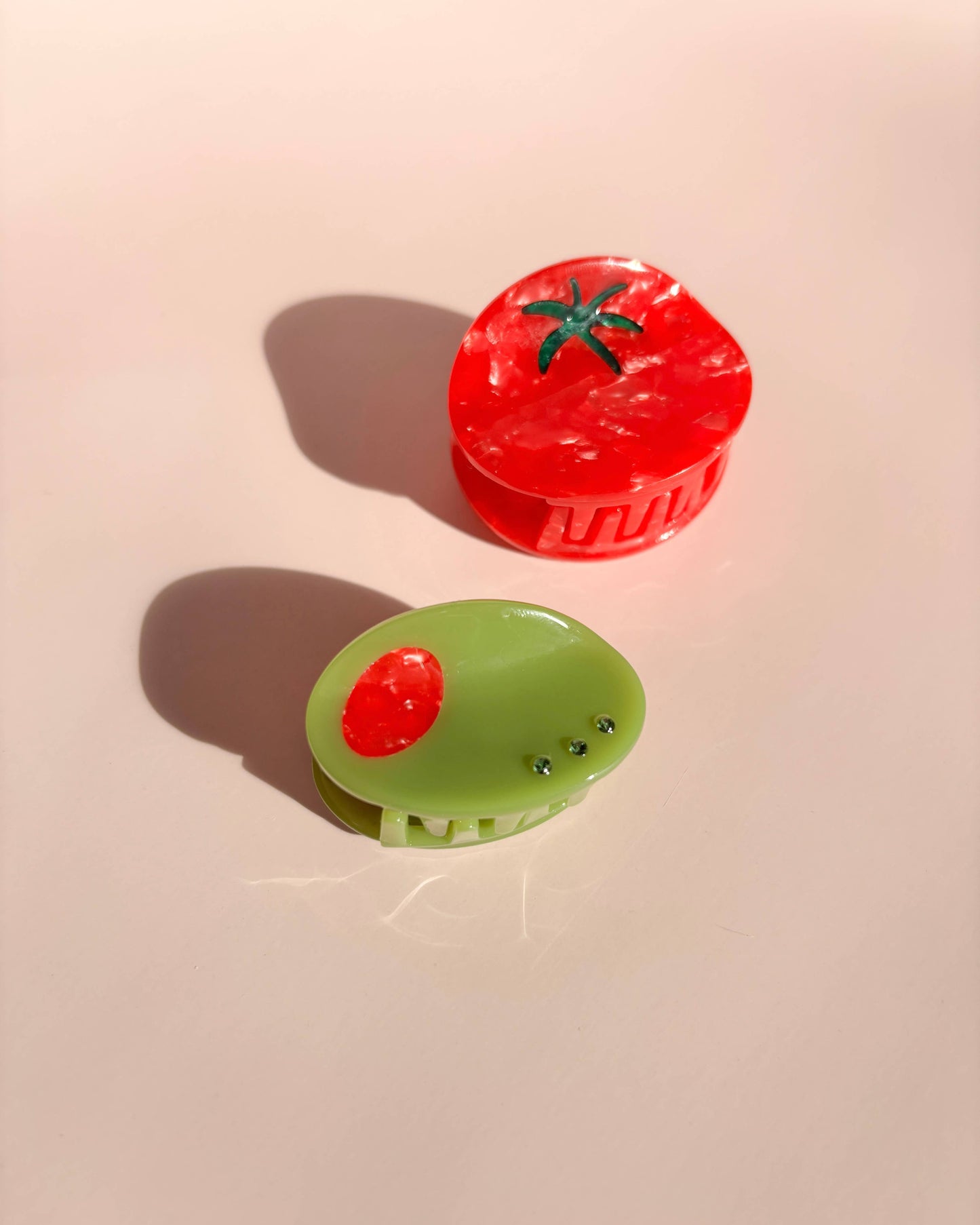 Two hair clips, one a tomato and one an olive, on a beige background.