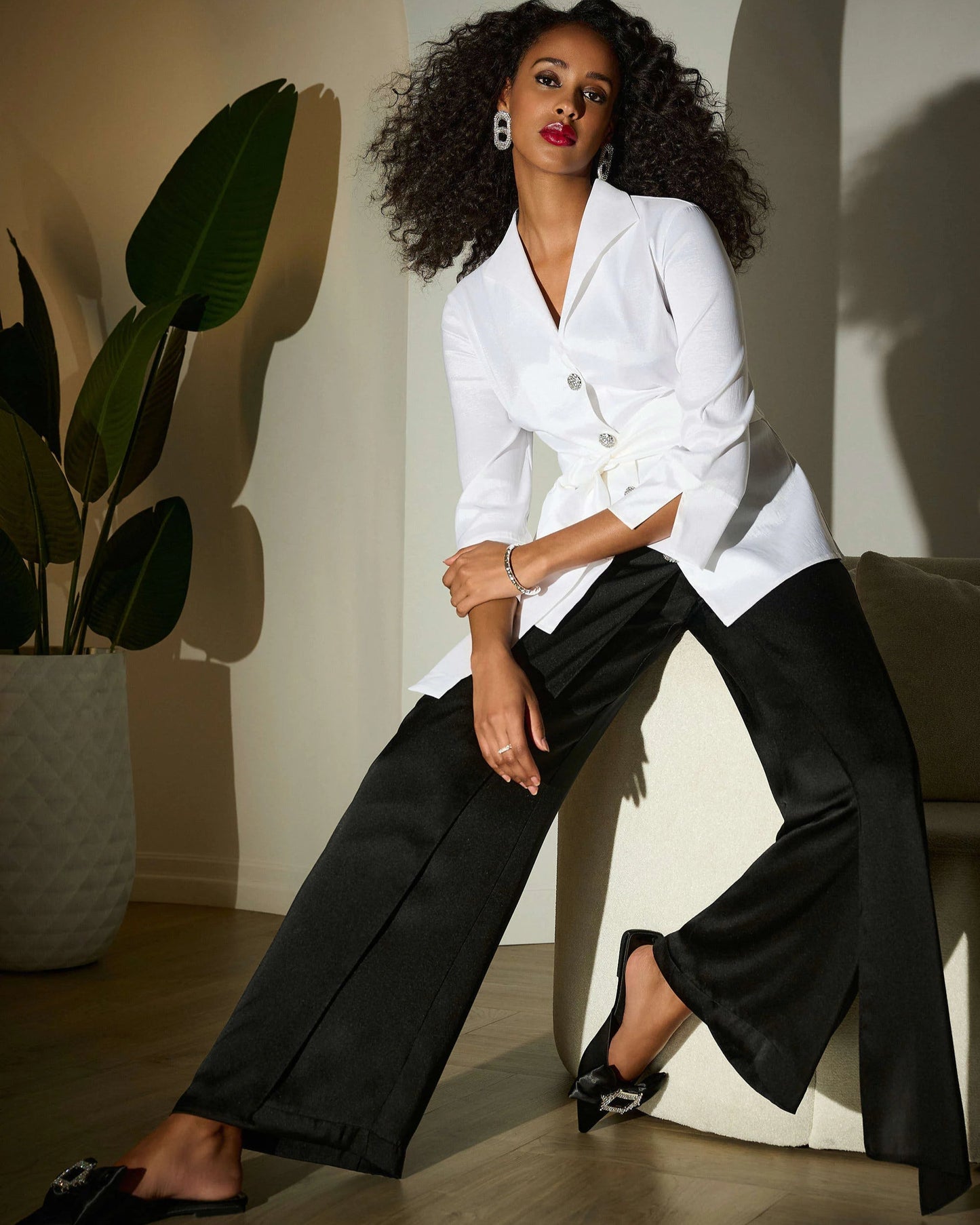 Woman in a white blazer and black pants sitting on a couch with a plant in the background
