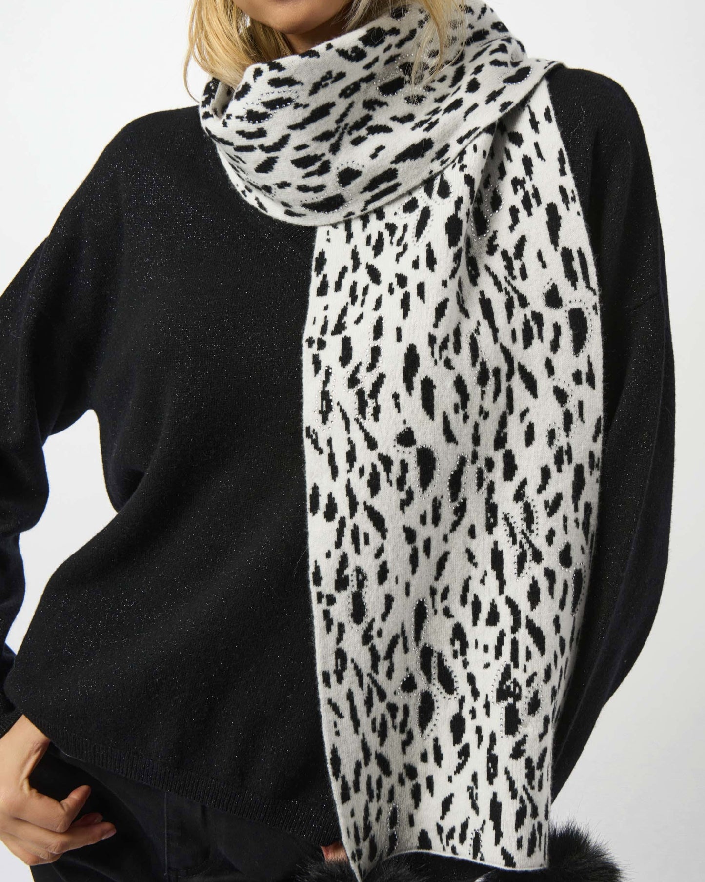 Person wearing a black sweater and a white scarf with black leopard print and pom-poms on a white background