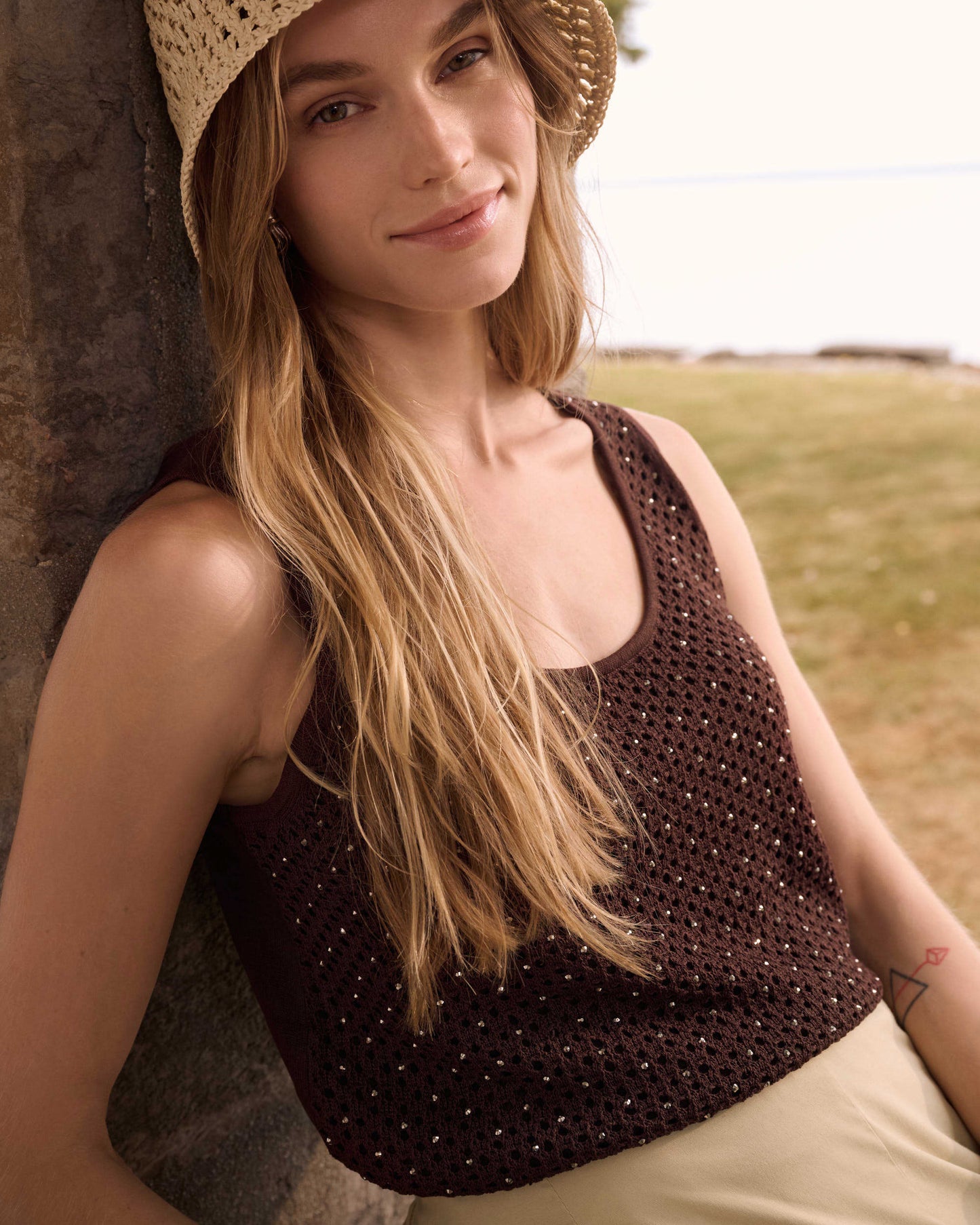 Woman wearing a straw hat and sleeveless top, leaning against a stone wall with a natural background.