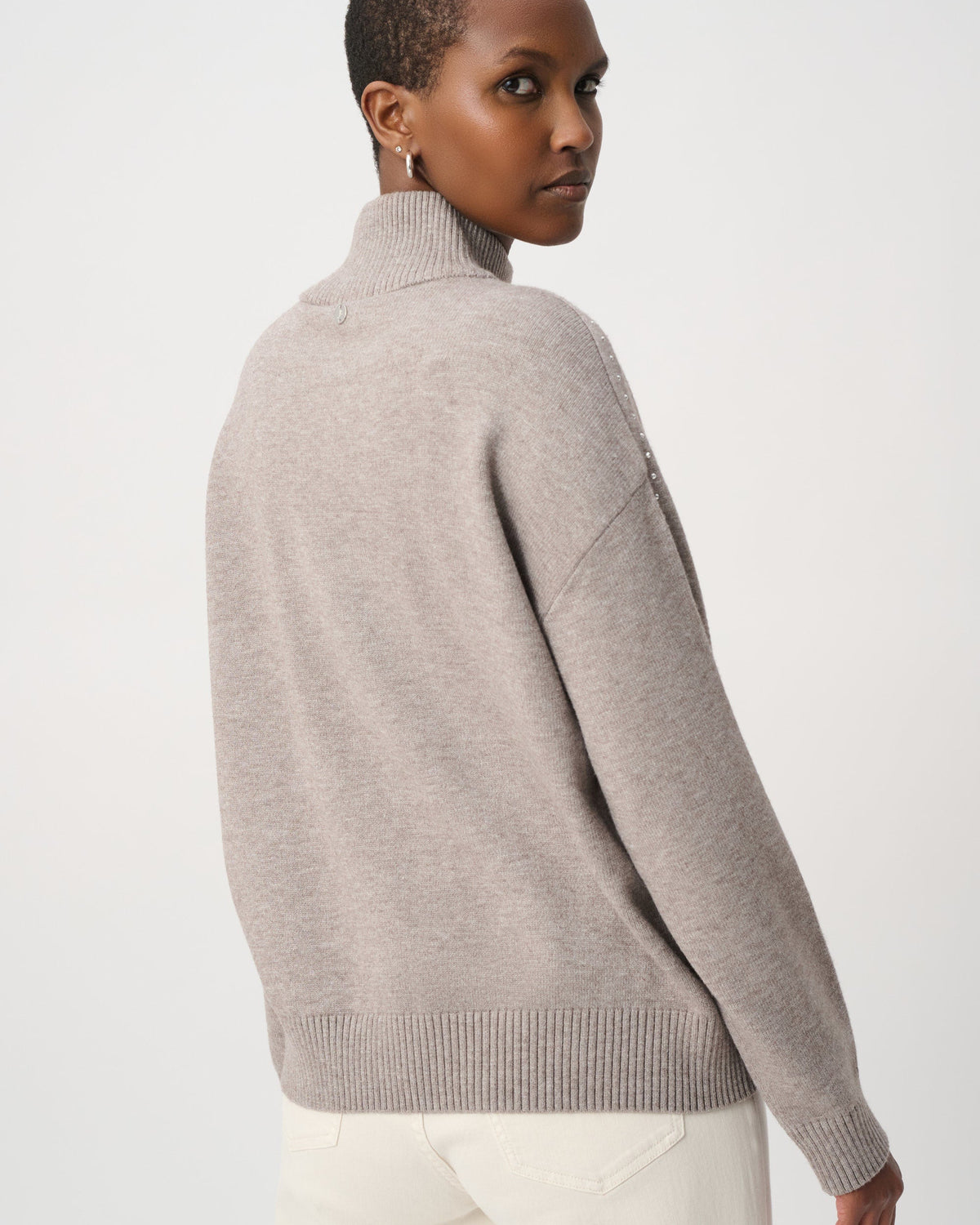 A model shown from the back wearing a light taupe knit mock neck sweater and white trousers, photographed against a clean white studio background.