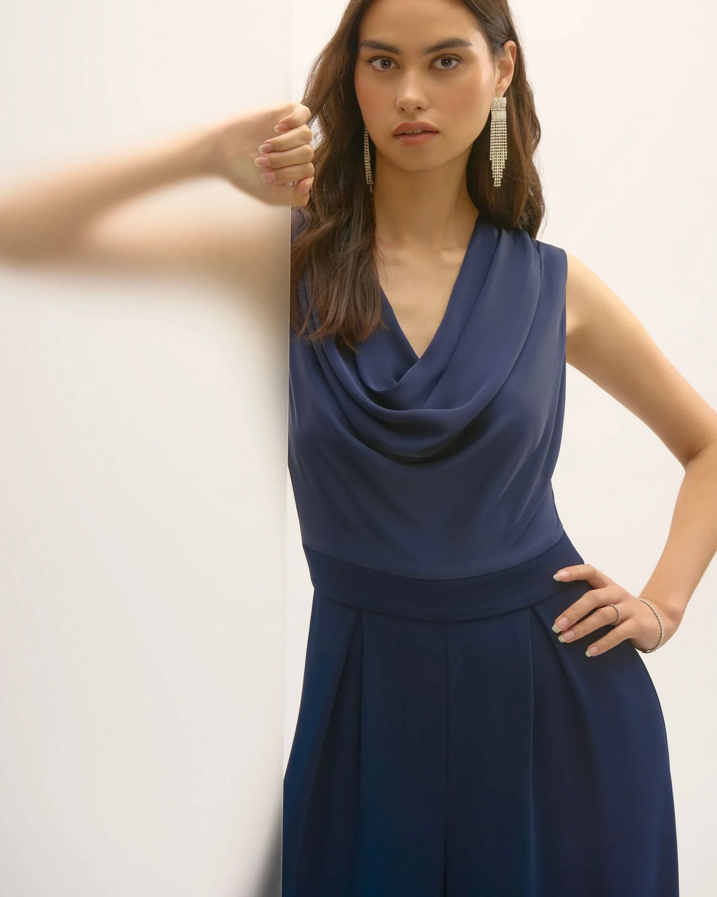 joseph ribkoff satin and crepe jumpsuit navy blue on model in front of white wall and glass wall, wearing dangly silver diamond earrings showing off top of jumpsuit