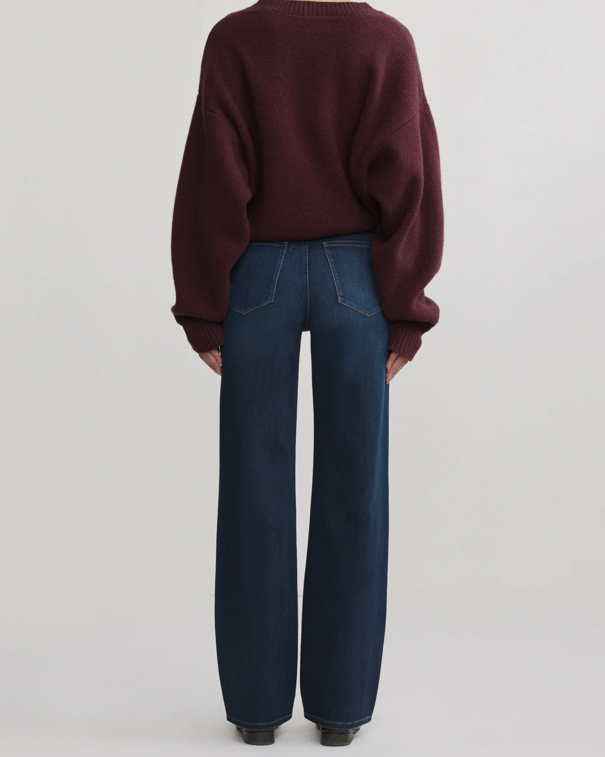 A model shown from the back wearing a dark burgundy knit sweater and dark blue wide-leg jeans against a light gray studio background.