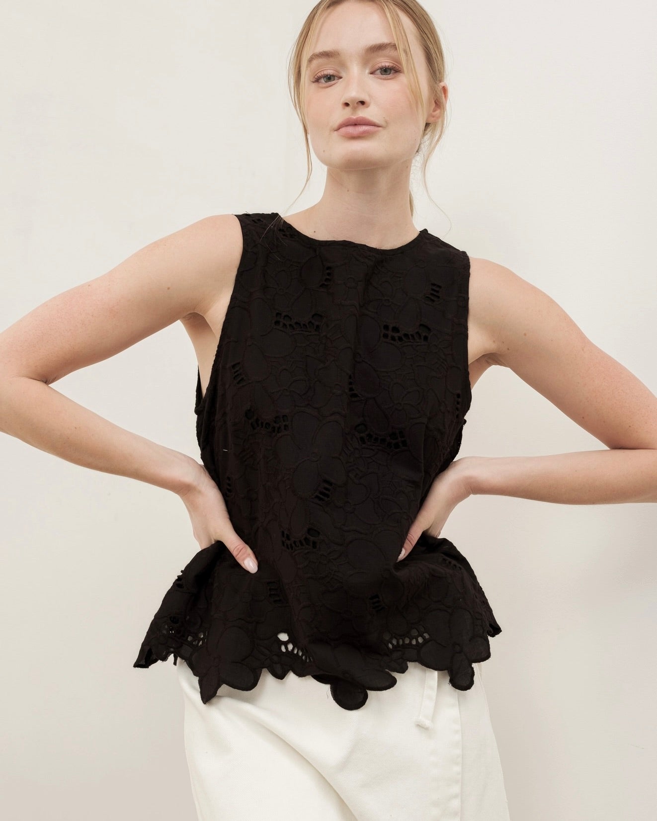 Modeling wearing the Aaron & Amber flower embroidered sleeveless top in black with hands on their hips in front of a white background
