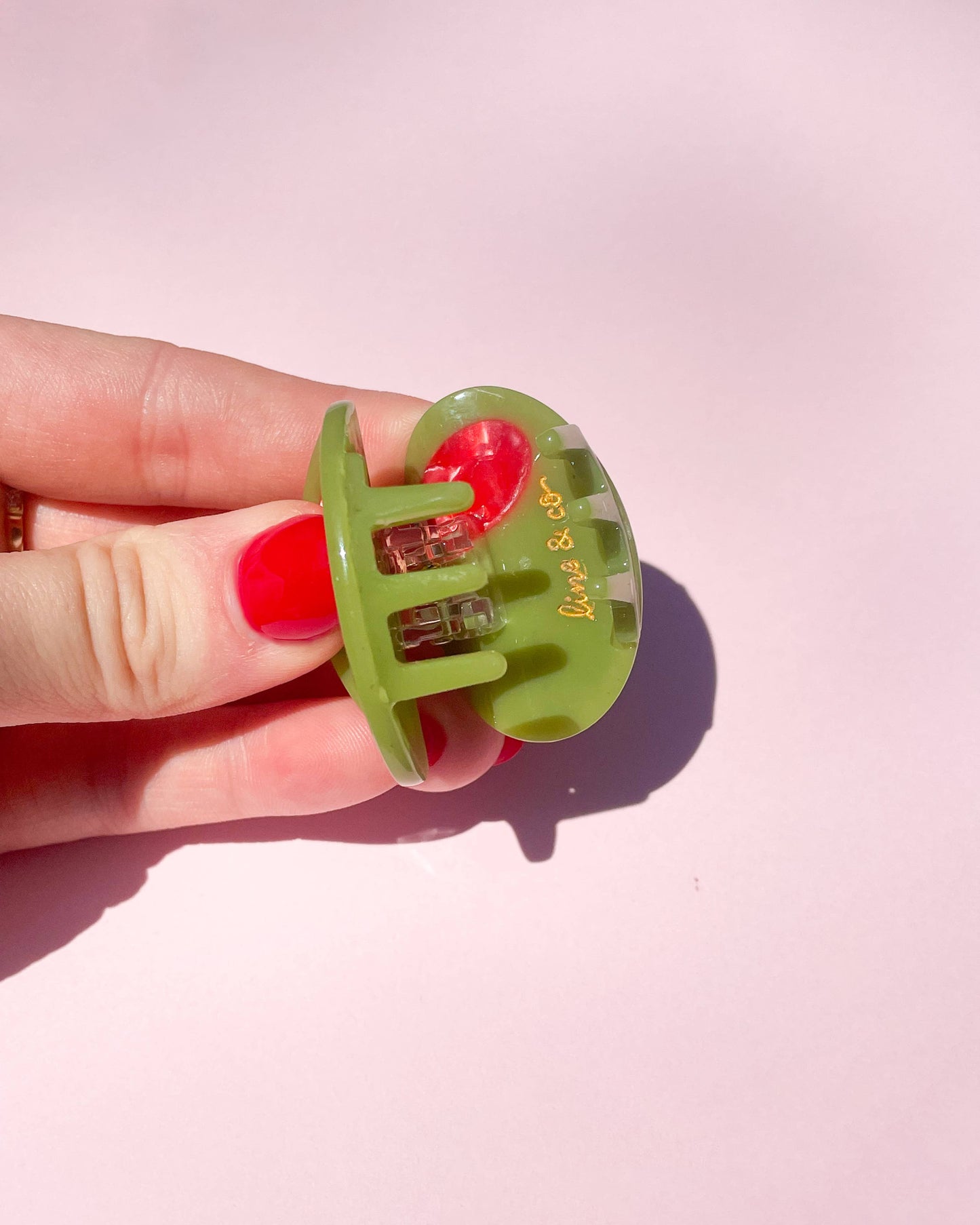 Green olive hair clip held by a hand against a pink background