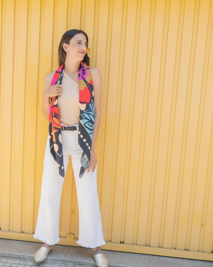 Woman wearing a colorful butterfly-patterned scarf and white pants and a black belt against a yellow wall.