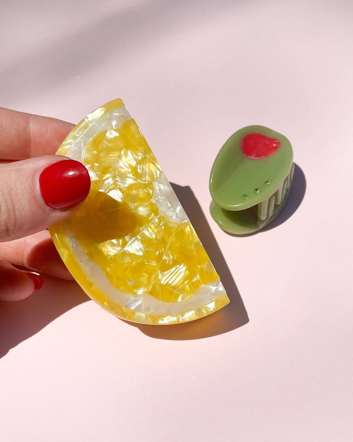 Hand holding a yellow lemon-shaped clip with a green olive-shaped clip on a pink background