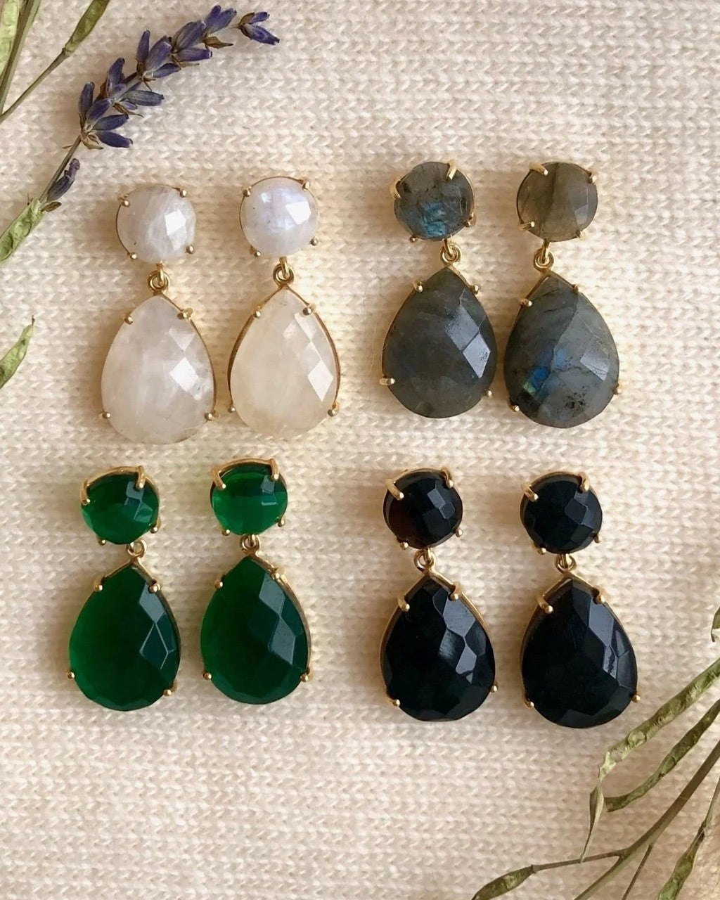 A collection of double drop earrings with gemstones, set in 14K gold vermeil, displayed on a fabric surface with lavender and greenery accents.