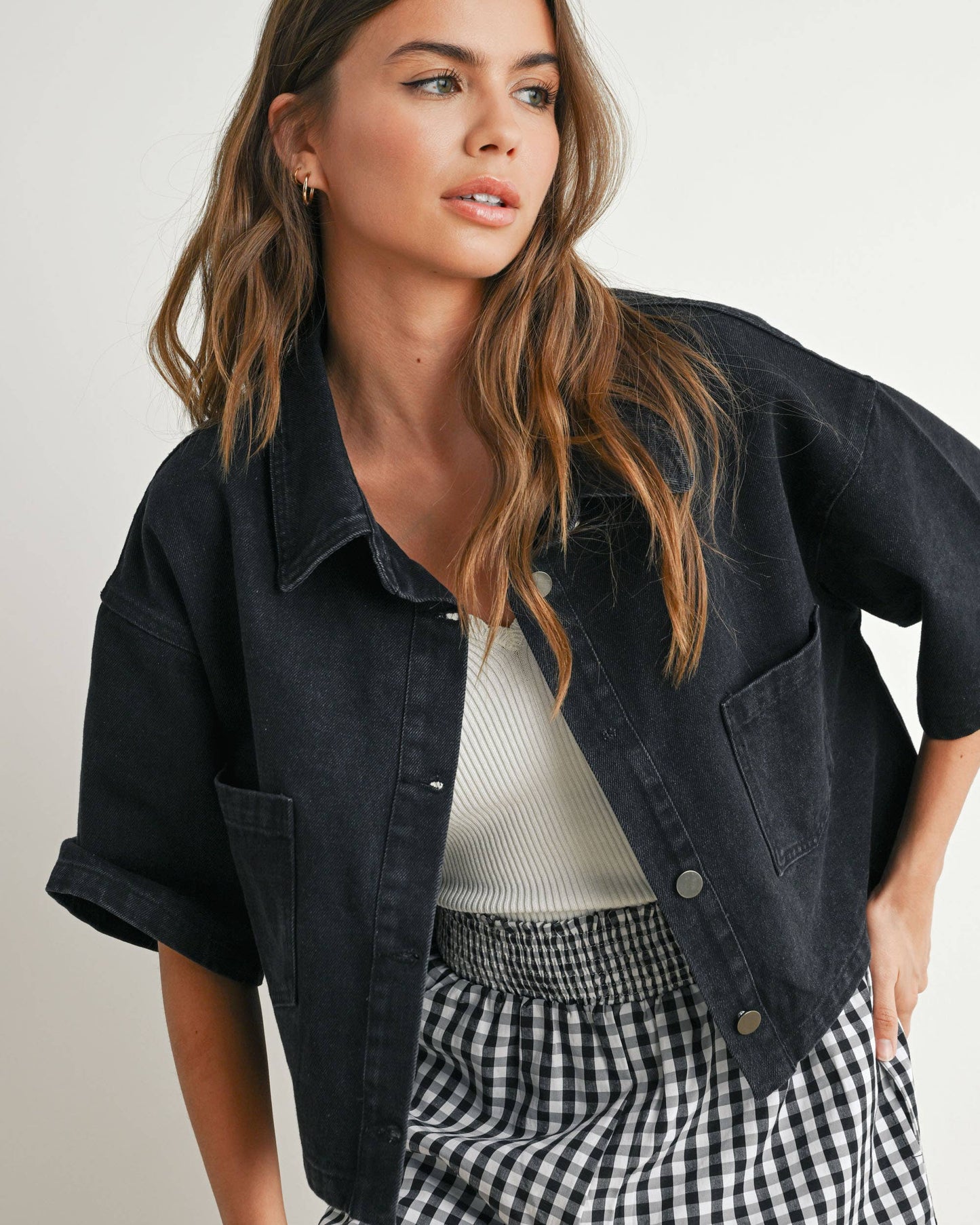 Woman wearing a black denim jacket, white top, and checkered pants on a plain background