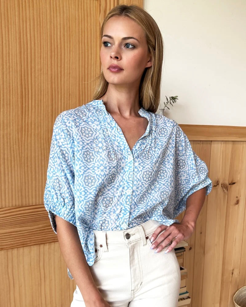 Model wearing Emerson Fry Mandarin Collar Top in Blue Geo Flower color wearing white pants standing in front of a brown wall