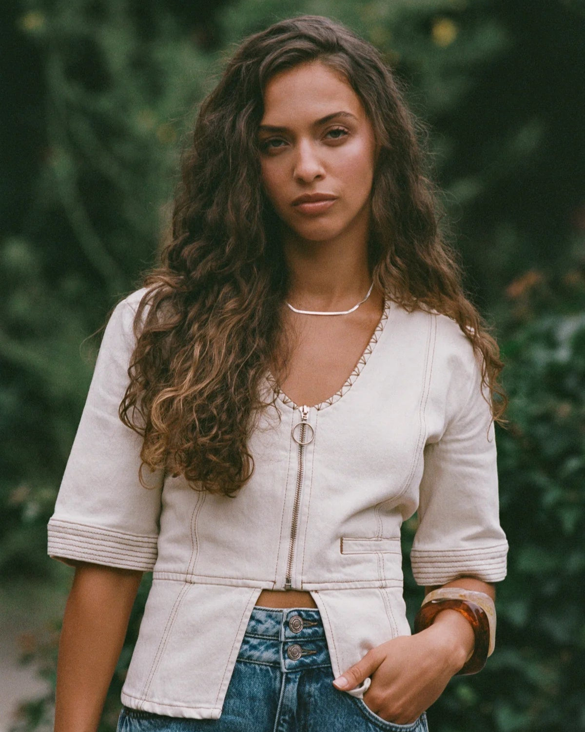 Woman wearing the lea top in ecru and blue jeans against a blurred natural background
