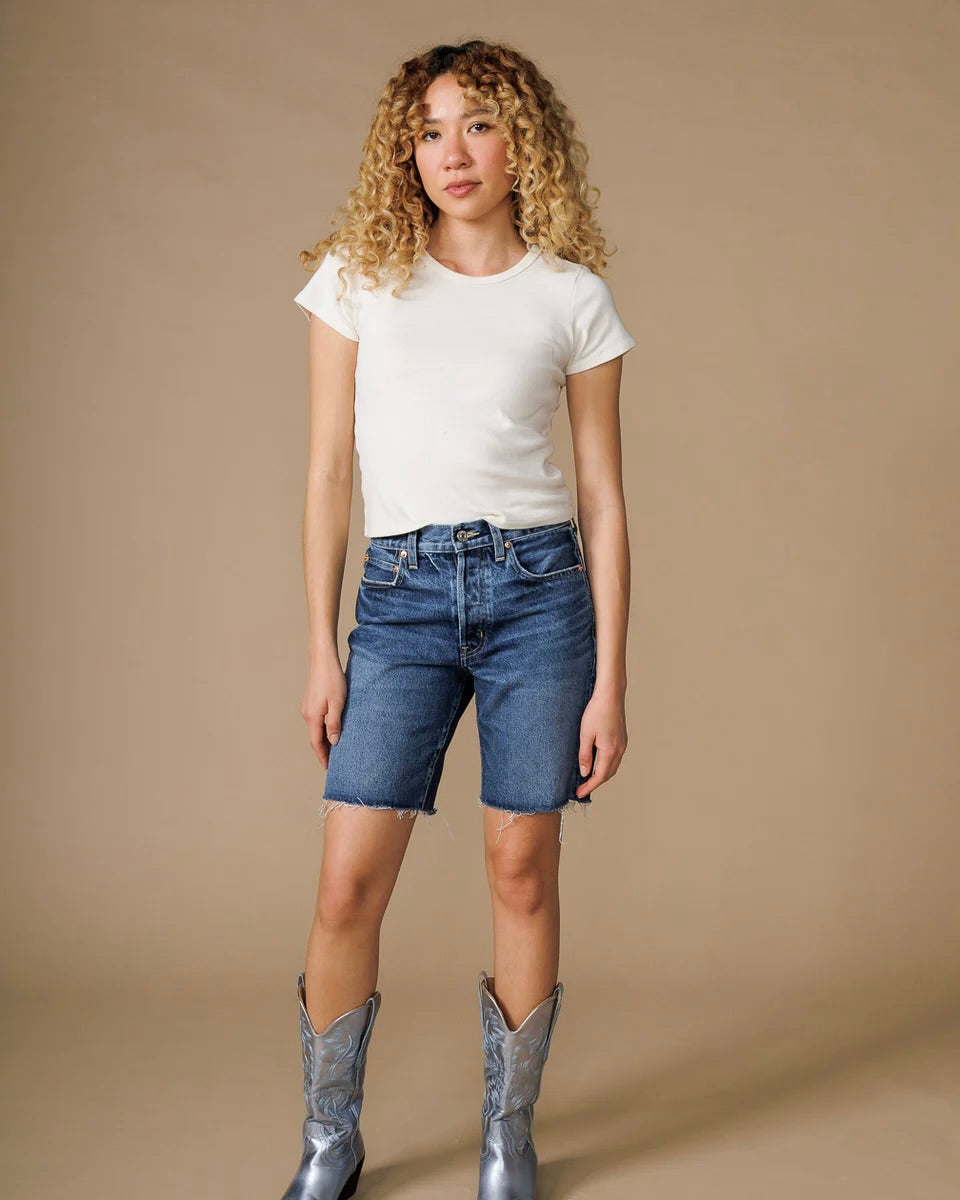 Model wearing the Edwin Denim Sawyer Bermuda short in peck with a white tee and cowboy boots standing in front of a neutral backdrop