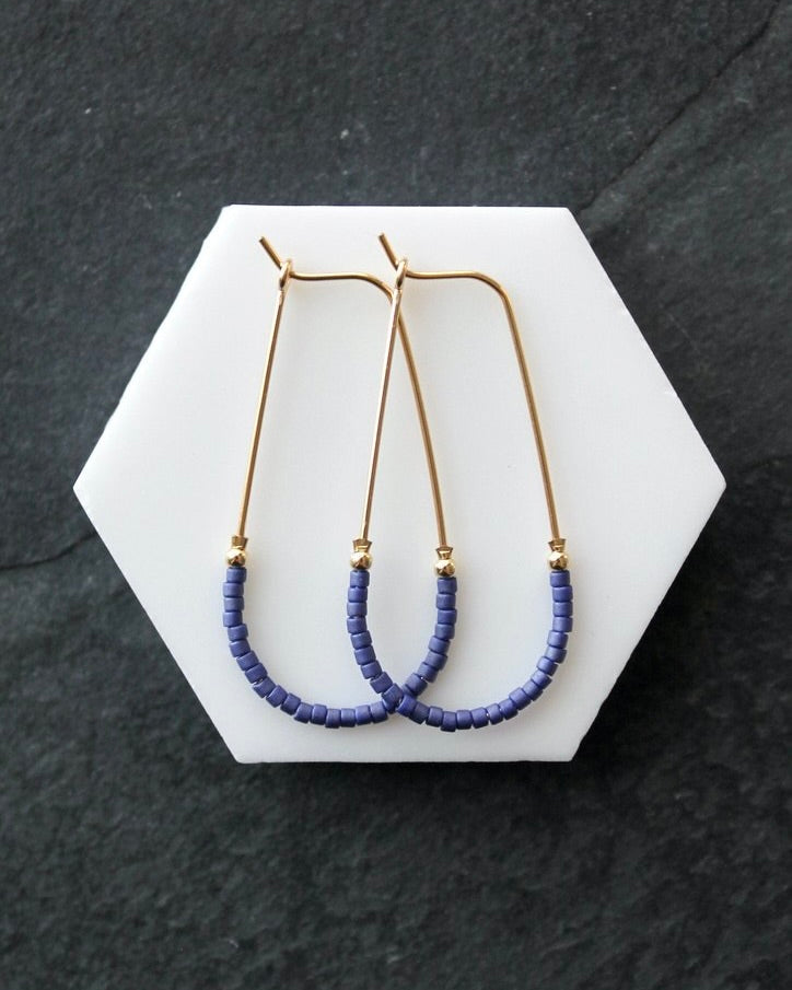 Close up photo of the Wild Balance Waterbury Hoop Earrings in gold with blue beads, laying against a white surface.