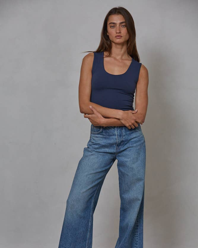Woman wearing a blue tank top and jeans standing against a plain background