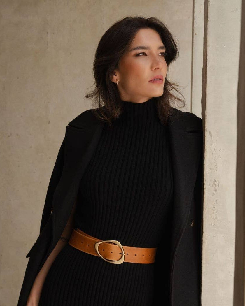 Woman wearing a black turtleneck dress with a brown belt against a neutral wall.