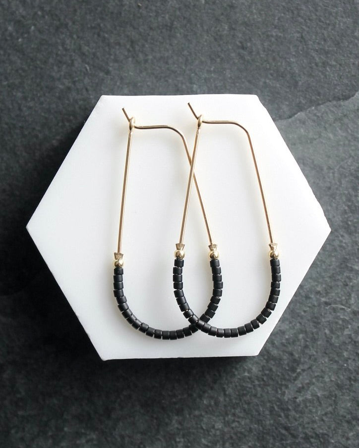 Close up photo of the Wild Balance Waterbury Hoop Earrings in gold with black beads, laying against a white surface.