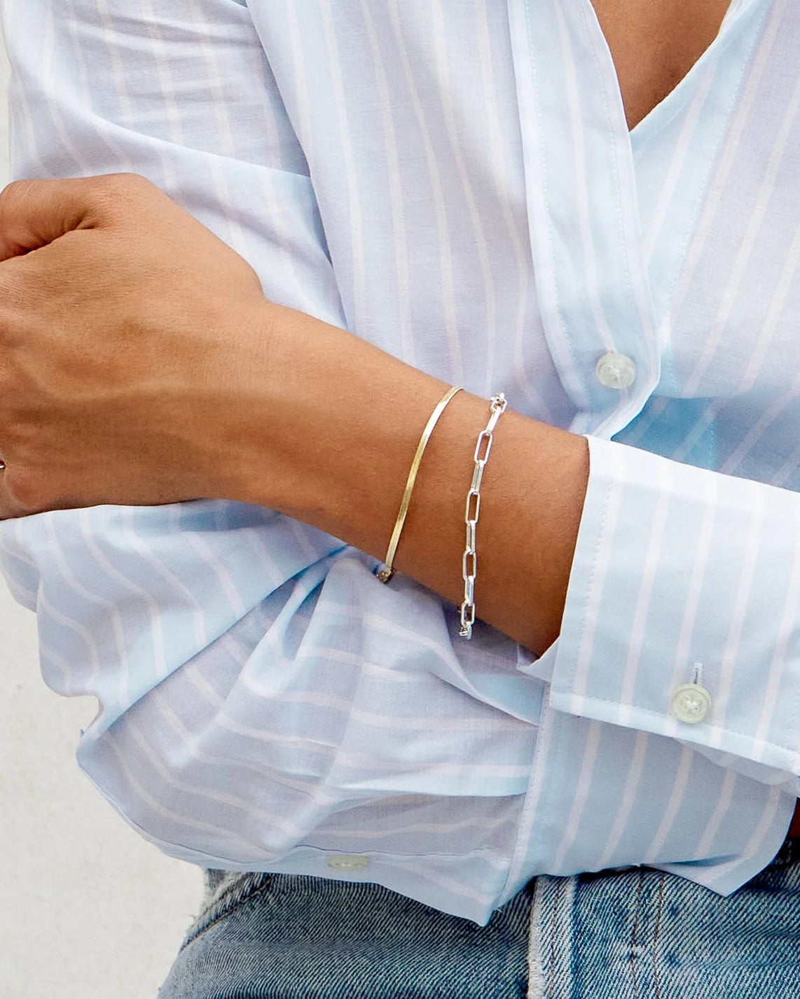 Model is wearing Gorjana Mini Venice Bracelet in gold and a simple silver chain bracelet and a blue button-up shirt