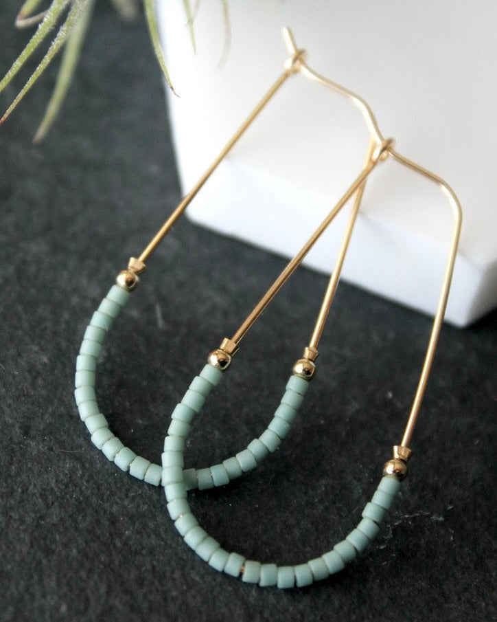 Close up photo of the Wild Balance Waterbury Hoop Earrings in gold with teal beads, laying against a gray and white background.