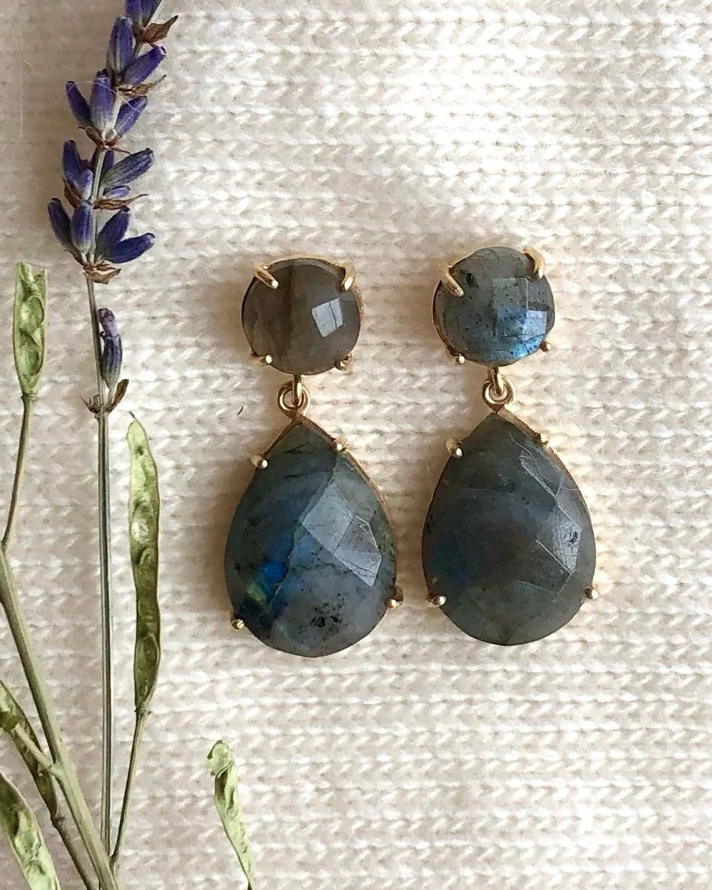 A pair of double drop earrings with gemstones, set in 14K gold vermeil, displayed on a fabric surface with lavender and greenery accents.