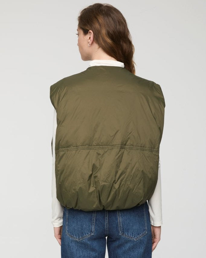 A woman shown from the back wearing an olive green puffer vest over a white collared shirt and blue jeans, against a plain light gray studio background.