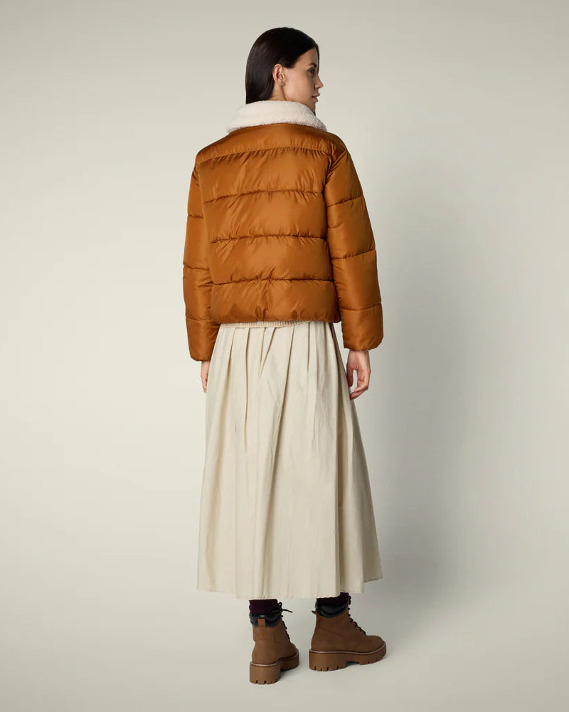 Woman wearing a brown puffer jacket and beige skirt on a plain background