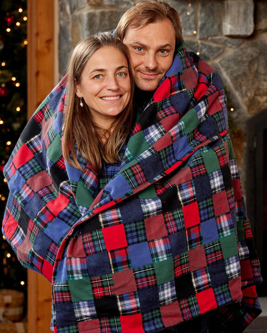 Two people wrapped in a large plaid blanket with a festive background