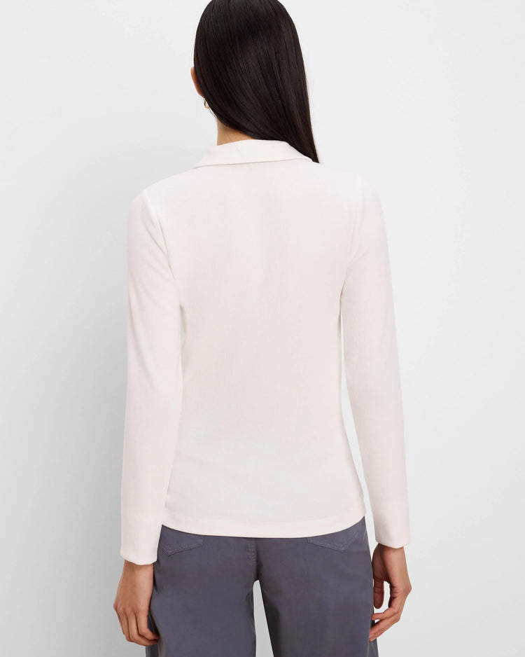 A woman viewed from the back wearing a light cream long-sleeve collared top and gray trousers, set against a plain light gray studio background.