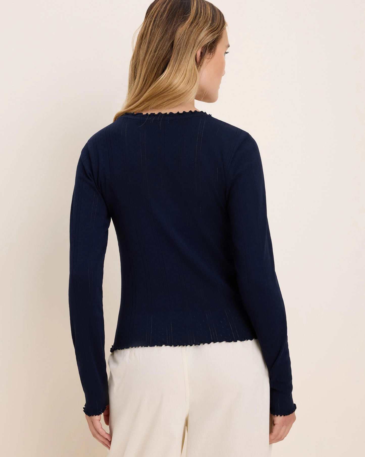 A woman shown from the back wearing a dark navy long-sleeve knit top with a scalloped neckline and hem, paired with cream pants, against a plain light beige studio background.