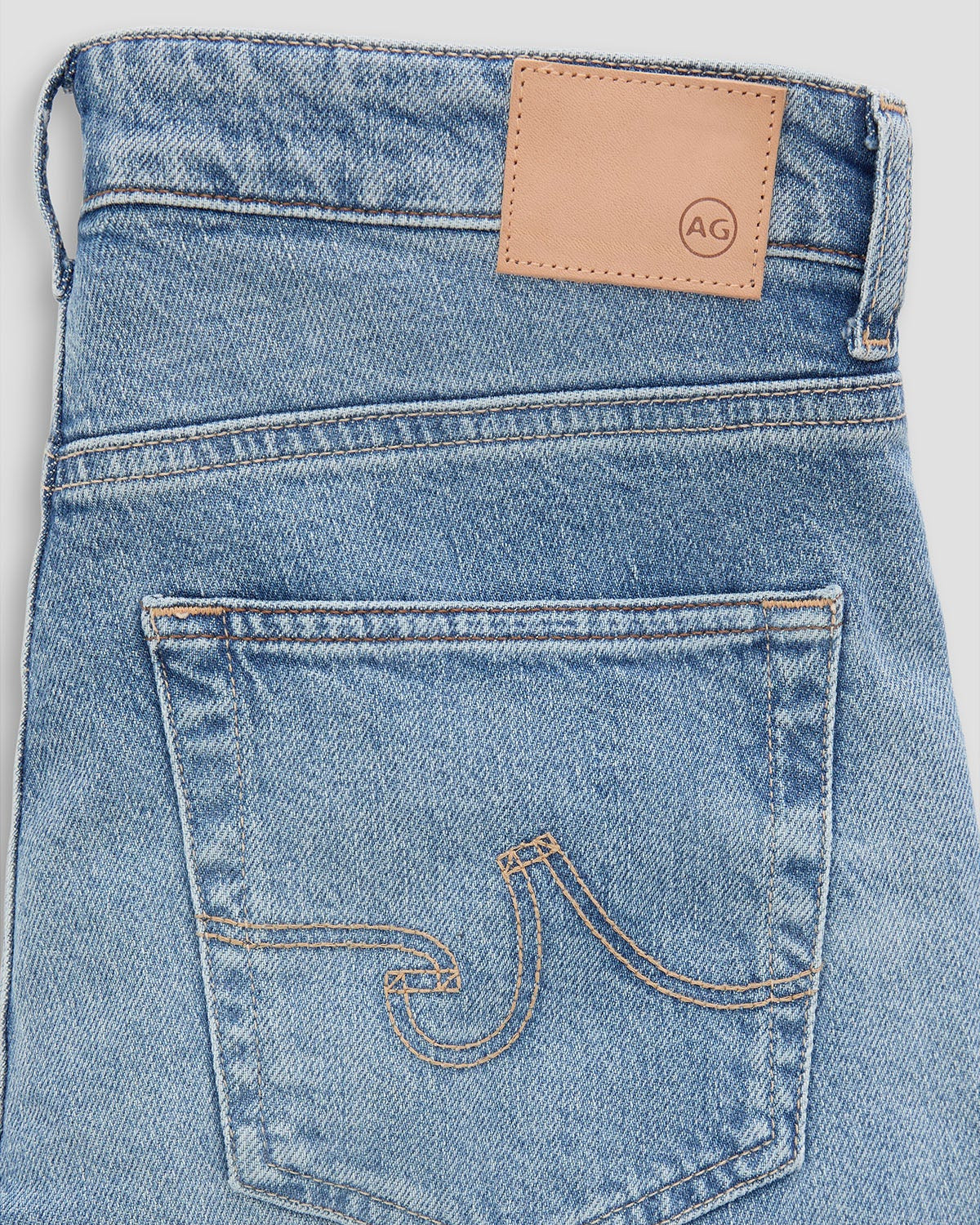 Close-up of the back waistband and pocket area of light-wash blue denim jeans, showing a tan leather patch with a circular AG logo and contrast stitching.