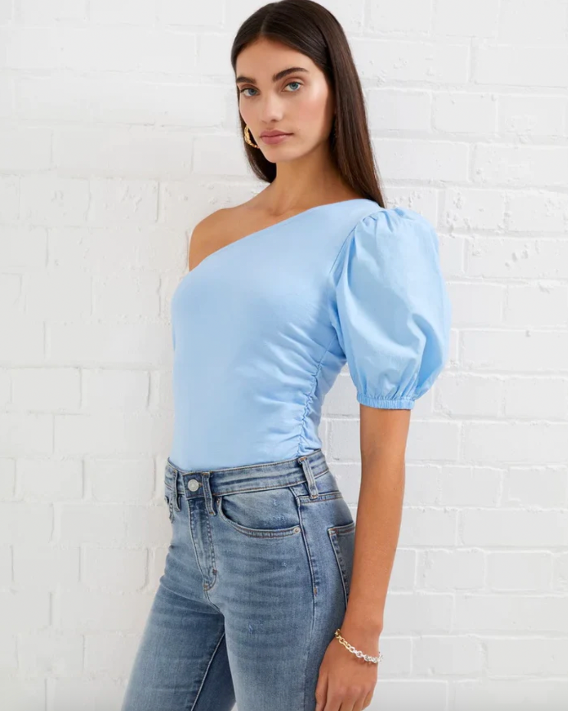 Model wearing French Connection Rosanna Cot mix 1 shoulder top in placid blue wearing jeans on a white brick background