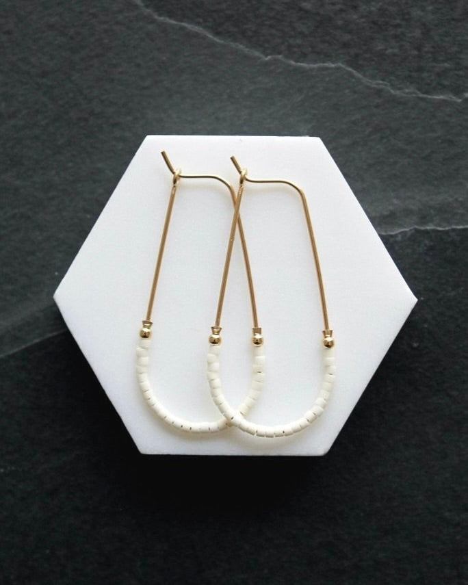 Close up photo of the Wild Balance Waterbury Hoop Earrings in gold with white beads, laying on a white surface.