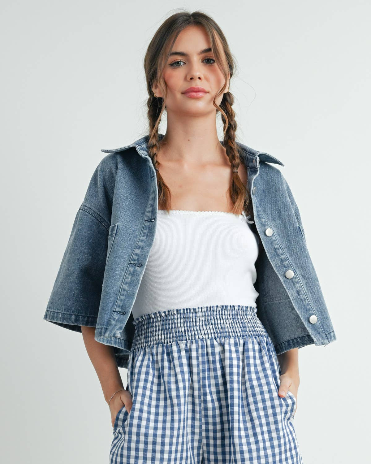 Woman wearing a denim jacket, white top, and blue checkered shorts on a plain background