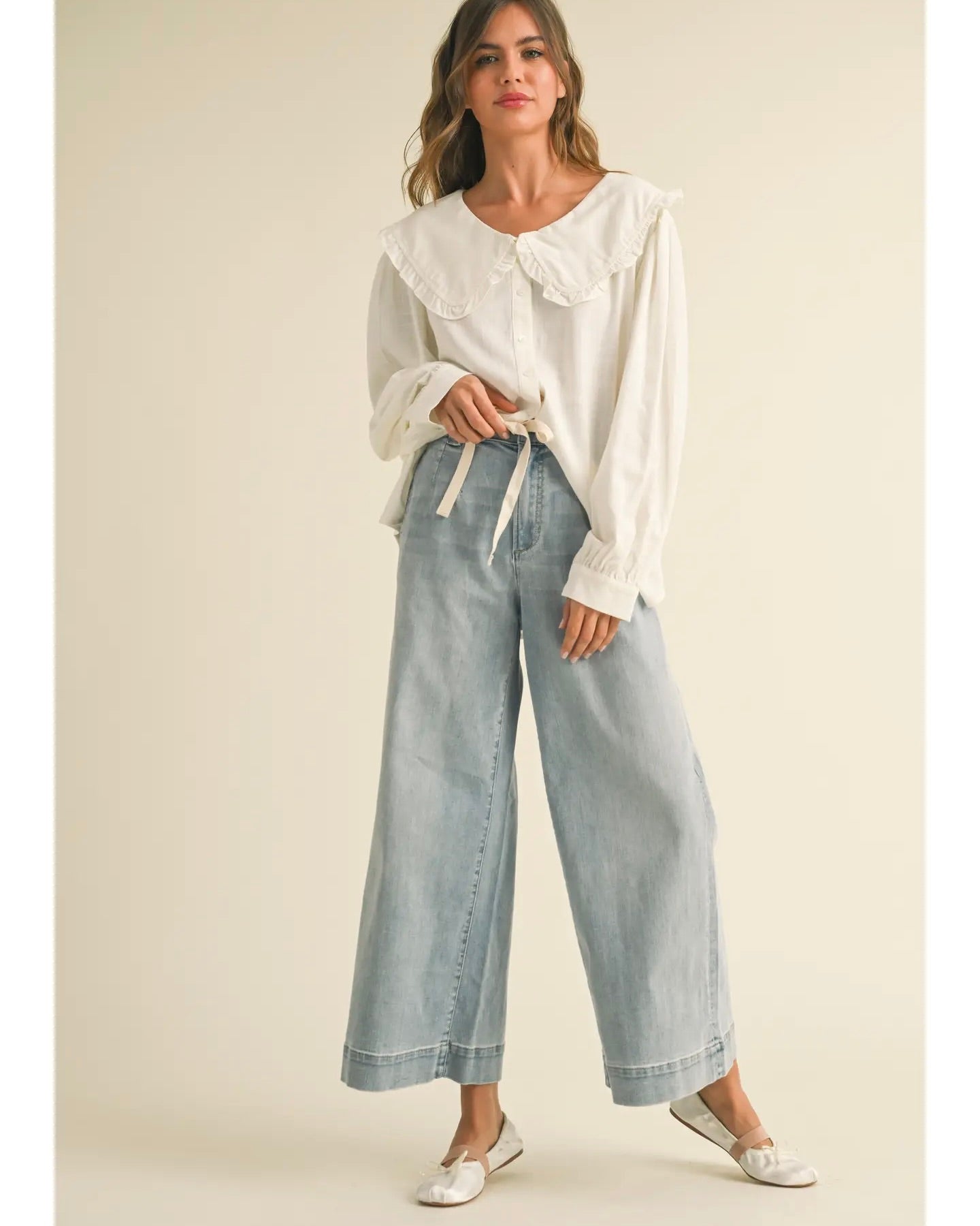 Model is wearing MIOU MUSE Washed Denim Tie Front Pants with a white blouse on a beige background.