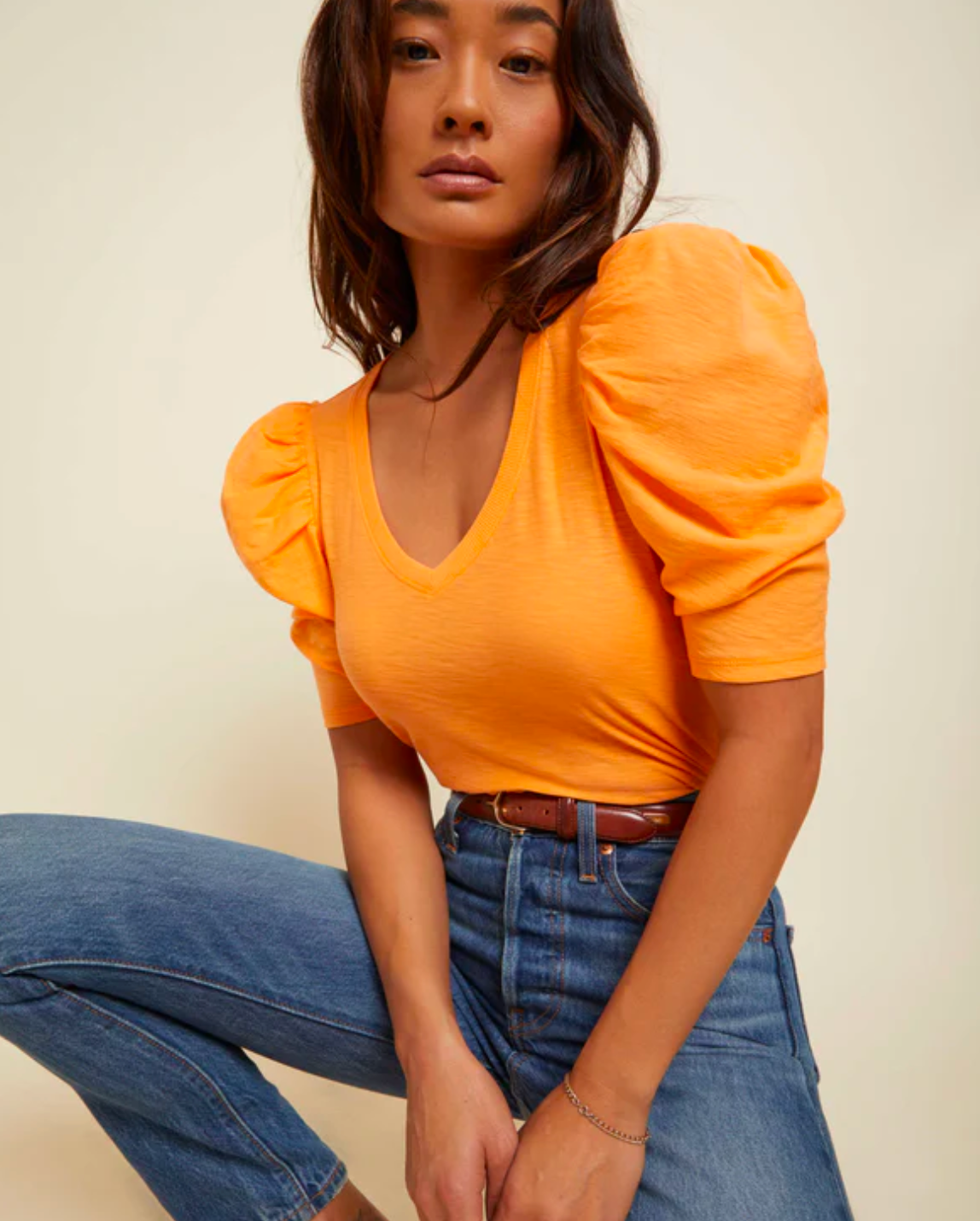 Model wearing Nation LTD Jillian Bold Shoulder V Neck In Tangerine color wearing jeans and a brown belt on an off white background