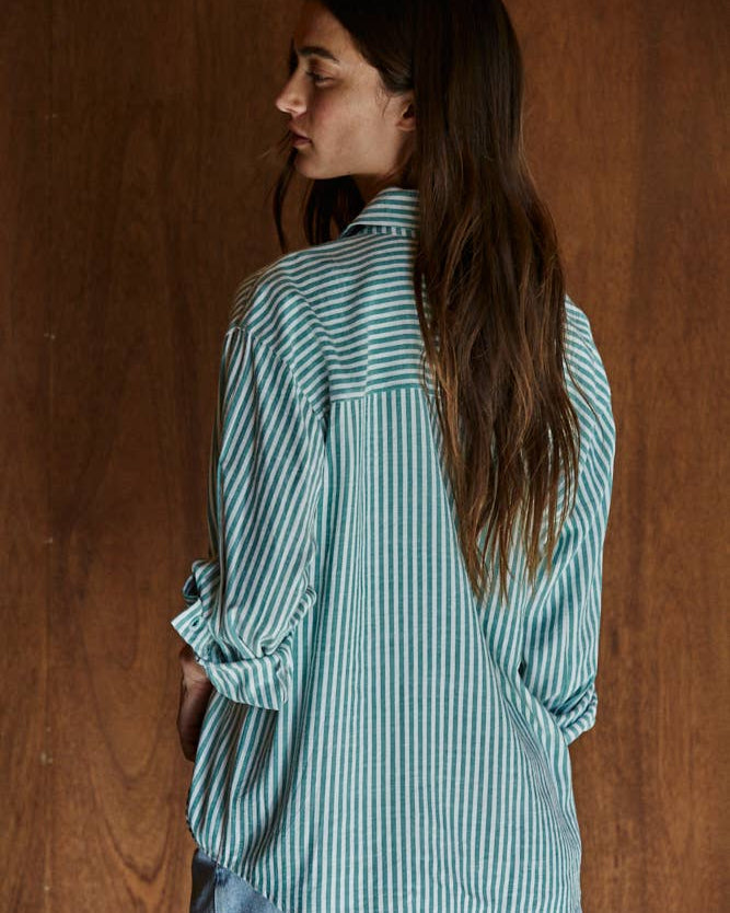 Back view of a woman wearing the By Together Woven Cotton Button Down in Green against a wooden background