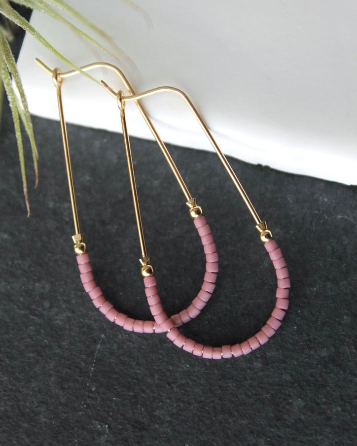Close up photo of the Wild Balance Waterbury Hoop Earrings in gold with pink beads, laying against a gray and white background.