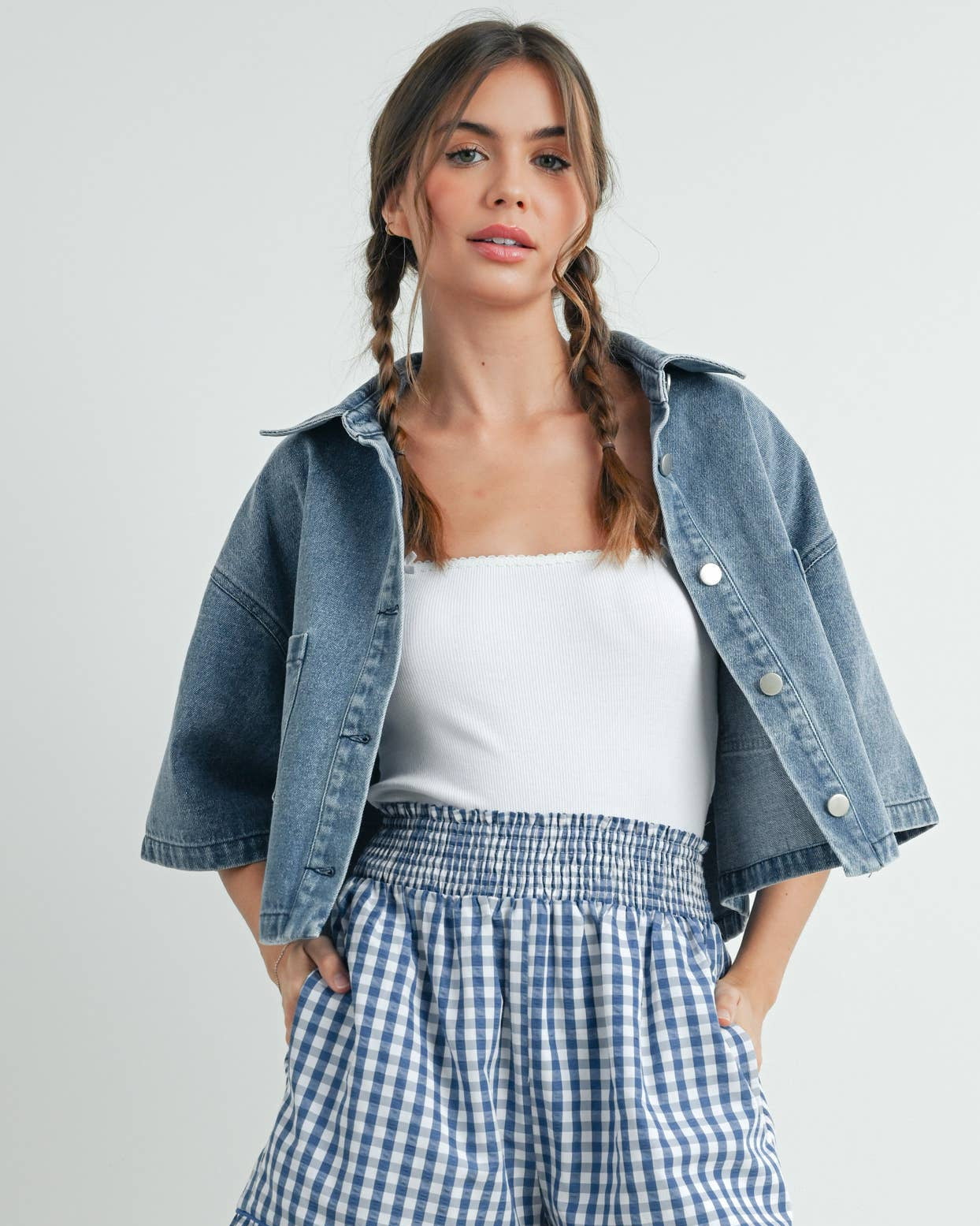 Woman wearing a denim jacket, white top, and blue checkered shorts on a light gray background