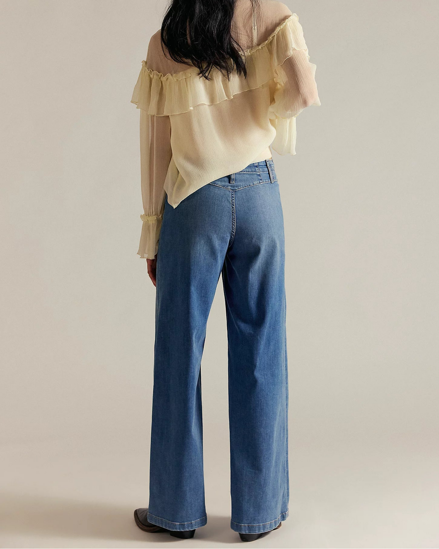Person wearing a beige layered blouse and blue jeans against a plain background