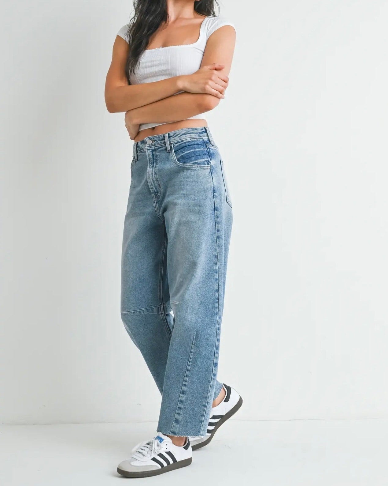 A person wearing a white tank top and blue barrel jeans with a mid-rise waist and 27-inch inseam, standing against a white background.