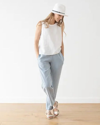 Model wearing LinenMe linen sleeveless Alice Blouse in white wearing blue trousers and a white hat on a white background standing on a wooden floor
