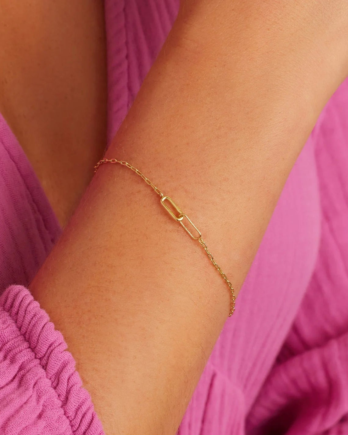 A person's wrist wearing a gold paperclip chain bracelet with two interlocking links at its center.