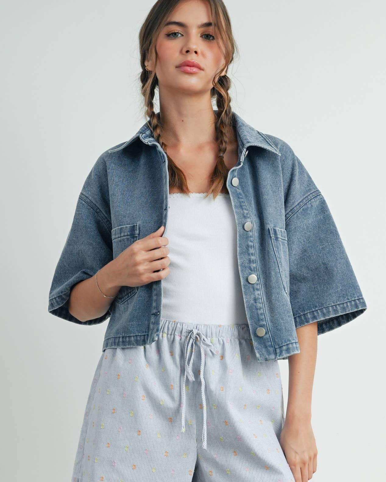 Woman wearing a denim jacket, white top, and light blue shorts on a plain background