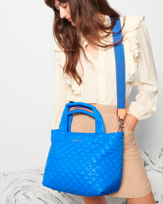 Model wearing MZ Wallace Small Metro tote deluxe bag in true blue color over the shoulder wearing a tan short skirt and an off white blouse on a white background