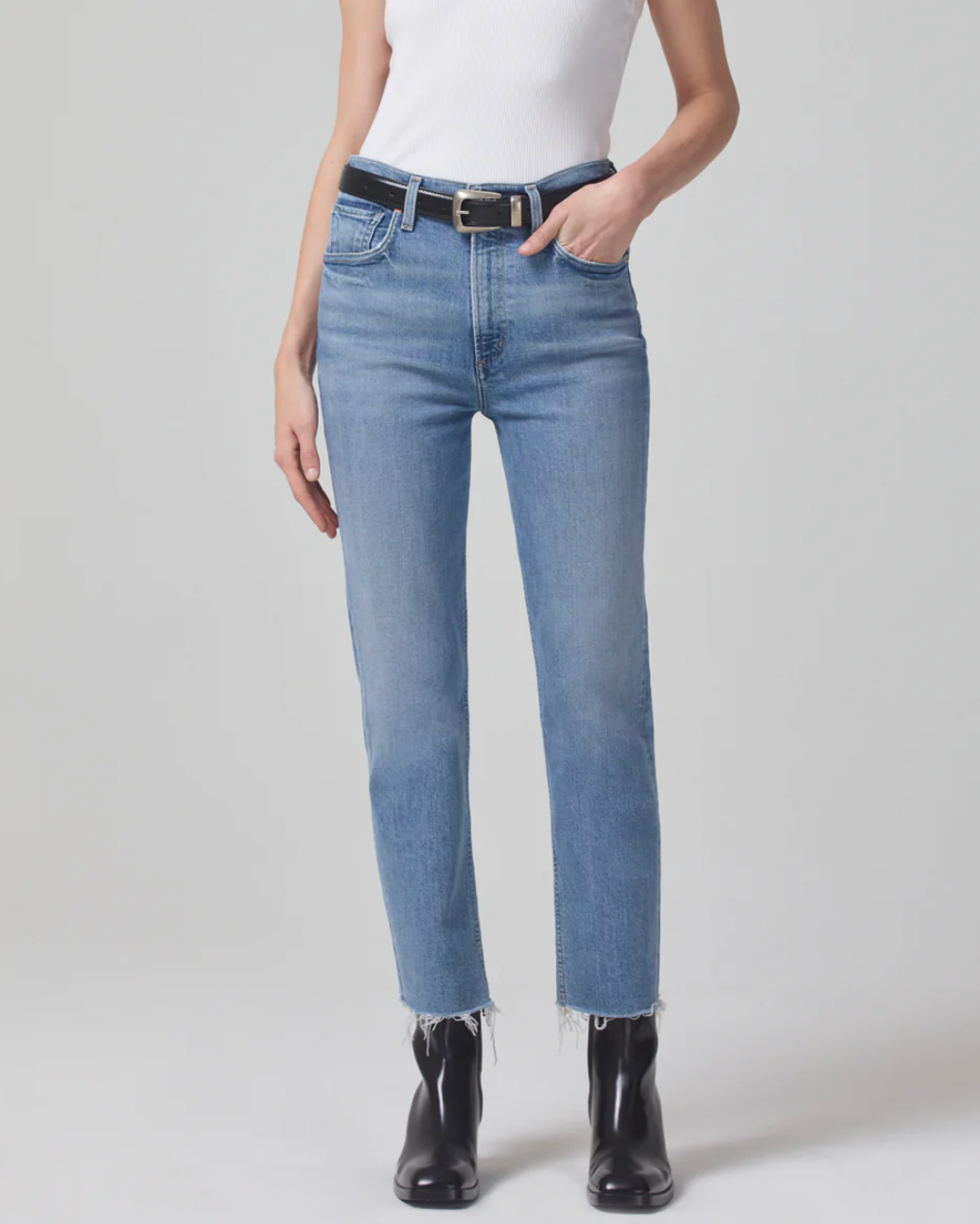 Model wearing Citizens of Humanity Daphne Crop High Rise stovepipe jean wearing black boots with a black belt and white tank top on a white background