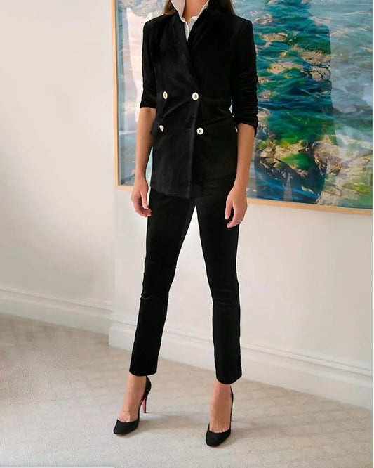 Model wearing Gretchen Scott Velvet Black Gripeless pants wearing black velvet blazer and black heels standing behind art 